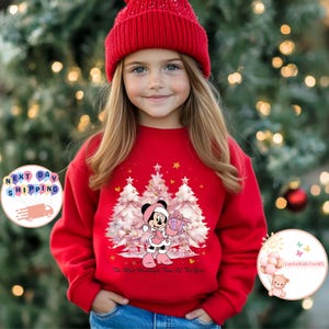May include: A red sweatshirt featuring a Minnie Mouse graphic holding a gift in front of Christmas trees. The text "The Most Wonderful Time Of The Year" is printed at the bottom. The girl is wearing a red beanie.