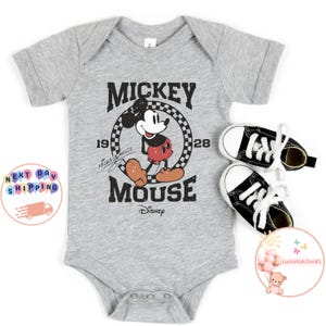K&ouml;nnte beinhalten: Ein grauer Kurzarm-Body mit einem Vintage-Mickey-Mouse-Motiv. Das Motiv zeigt Mickey Mouse in einem rot-schwarzen Outfit, der auf einem karierten Kreis steht, mit dem Text "Mickey Mouse" und "1928" um den Kreis herum. Der Text "Mickey Mouse" ist in einer handschriftlichen Schriftart geschrieben und der Text "Disney" befindet sich unter dem Motiv.