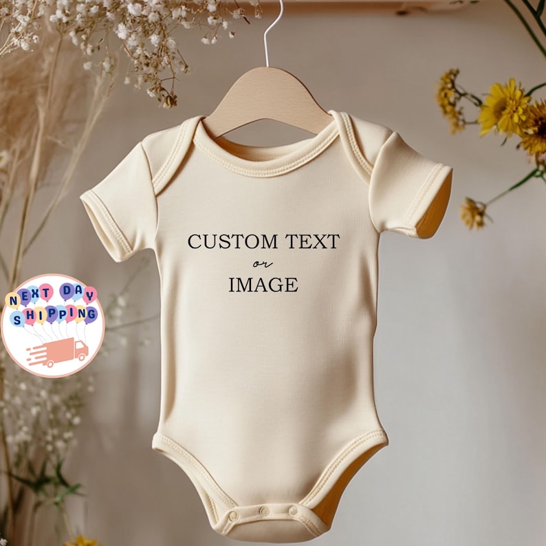 May include: A light beige baby onesie with short sleeves hangs on a wooden hanger. The onesie has the text "CUSTOM TEXT or IMAGE" printed in black. The background includes flowers and a "NEXT DAY SHIPPING" graphic.