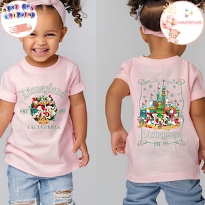 May include: Pink t-shirt with a Christmas-themed Disneyland graphic featuring Mickey Mouse and other Disney characters. The front of the shirt says "Disneyland EST 1955 CALIFORNIA". The back of the shirt features a Christmas-themed Disneyland castle with Mickey Mouse and other Disney characters. The back of the shirt says "Disneyland EST 1955".