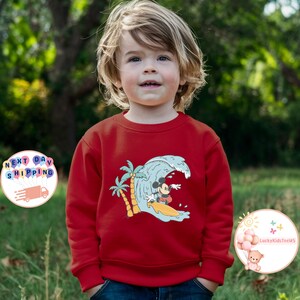 Puede incluir: Sudadera roja de manga larga con un gr&aacute;fico de Mickey Mouse surfeando en una ola con palmeras en el fondo.