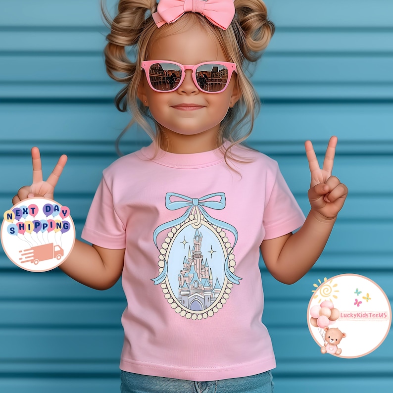 Puede incluir: Una camiseta rosa con un gr&aacute;fico de un castillo azul y rosa en un marco de perlas. El castillo est&aacute; rodeado de estrellas y un lazo azul. La camiseta est&aacute; puesta en una ni&ntilde;a que lleva gafas de sol rosas.