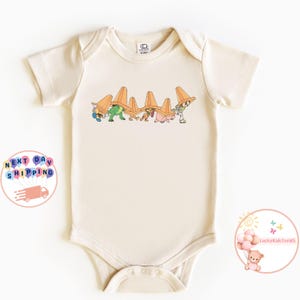 May include: Cream-colored baby bodysuit featuring a colorful graphic of cartoon characters wearing orange traffic cones. The design includes various characters in different poses. The bodysuit is made of soft fabric and has short sleeves.