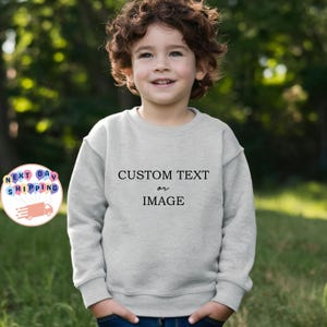 May include: A light gray sweatshirt with the text "CUSTOM TEXT or IMAGE" in black. The sweatshirt is worn by a child with curly brown hair and blue jeans. The background is blurred green foliage.