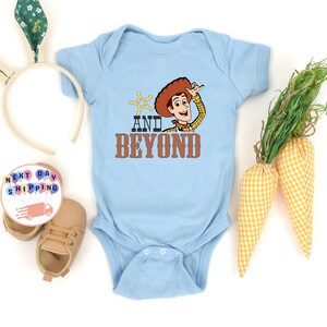 May include: A light blue baby bodysuit featuring a cartoon image of Woody from Toy Story with the words "AND BEYOND". The bodysuit has a snap closure. The image also includes a pair of baby shoes, a bunny ear headband, and decorative carrots.