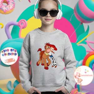 May include: A light gray sweatshirt featuring a cartoon image of a cowgirl hugging a brown horse. The cowgirl wears a red hat, white shirt, and blue jeans with a cow print. The sweatshirt has a crew neck and long sleeves.