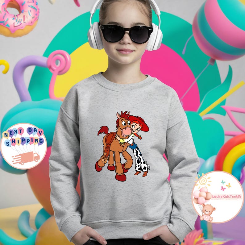 May include: A light gray sweatshirt featuring a cartoon image of a cowgirl hugging a brown horse. The cowgirl wears a red hat, white shirt, and blue jeans with a cow print. The sweatshirt has a crew neck and long sleeves.
