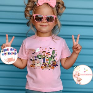 Puede incluir: Una camiseta rosa claro con un gr&aacute;fico de personajes de Toy Story con tem&aacute;tica de Halloween. La camiseta la lleva un ni&ntilde;o peque&ntilde;o que lleva gafas de sol rosas y un lazo rosa.