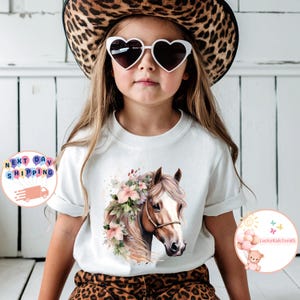 May include: White t-shirt featuring a watercolor illustration of a horse's head adorned with flowers. The horse has a brown coat and a white blaze. The model is wearing heart-shaped sunglasses and a leopard print hat.