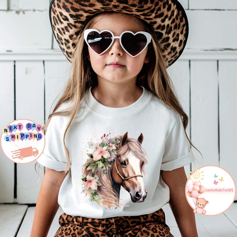 May include: White t-shirt featuring a watercolor illustration of a horse's head adorned with flowers. The horse has a brown coat and a white blaze. The model is wearing heart-shaped sunglasses and a leopard print hat.