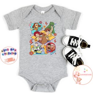 Op de afbeelding: Een grijze babybody met een kleurrijke grafische print van personages uit de Toy Story-franchise. De personages zijn Woody, Buzz Lightyear, Jessie, Rex en Mr. Potato Head. De grafiek is omringd door kleurrijke geometrische vormen en bliksemschichten.