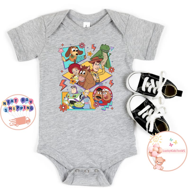 Op de afbeelding: Een grijze babybody met een kleurrijke grafische print van personages uit de Toy Story-franchise. De personages zijn Woody, Buzz Lightyear, Jessie, Rex en Mr. Potato Head. De grafiek is omringd door kleurrijke geometrische vormen en bliksemschichten.