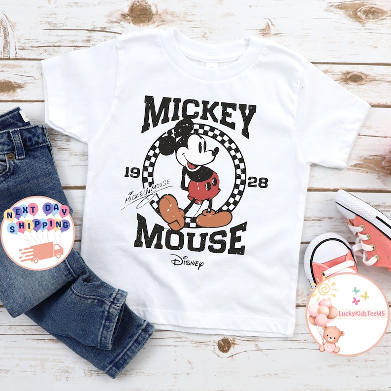 K&ouml;nnte beinhalten: Wei&szlig;es T-Shirt mit einem Vintage-Mickey-Mouse-Motiv. Das Motiv zeigt Mickey Mouse in einem schwarz-wei&szlig; karierten Kreis mit dem Text "Mickey Mouse" dar&uuml;ber und "1928" darunter. Der Text "Disney" befindet sich unter dem Motiv.