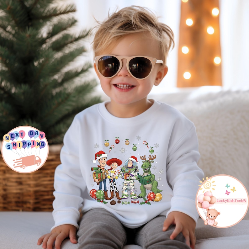 May include: A white sweatshirt with a Christmas-themed Toy Story graphic. The design features Woody, Jessie, Buzz Lightyear, and a green dinosaur, all wearing festive hats. The sweatshirt is worn by a child.