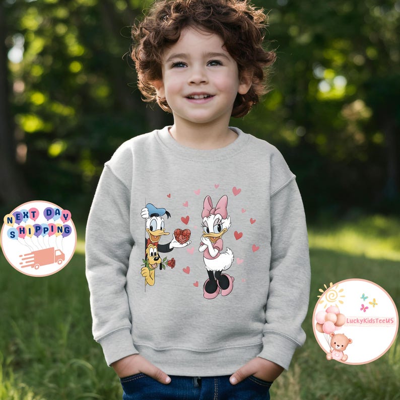 May include: A light gray sweatshirt featuring Donald Duck presenting a heart-shaped gift to Daisy Duck, surrounded by pink hearts. The design includes Pluto and roses. The sweatshirt is worn by a child.