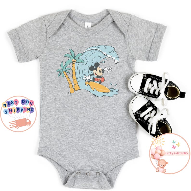 Puede incluir: Un body gris de manga corta con un gr&aacute;fico de Mickey Mouse surfeando en una ola con palmeras en el fondo.