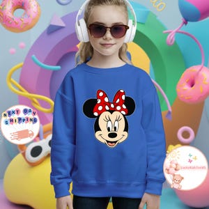 May include: A blue sweatshirt with a Minnie Mouse design, featuring a red polka dot bow. The image shows a young person wearing the sweatshirt, headphones, and sunglasses. The background includes colourful shapes and donuts.