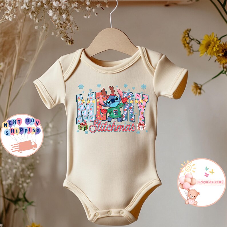 Puede incluir: Un body de beb&eacute; color crema con un gr&aacute;fico festivo de "Merry Stitchmas". El dise&ntilde;o presenta al personaje Stitch con cuernos de reno y sosteniendo luces navide&ntilde;as. El body est&aacute; colgado de una percha de madera.