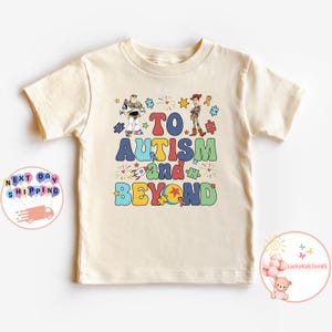 Puede incluir: Una camiseta beige de manga corta con un gr&aacute;fico colorido de los personajes Buzz Lightyear y Woody de la franquicia Toy Story. El gr&aacute;fico incluye el texto "To Autism and Beyond" con piezas de rompecabezas, estrellas y fuegos artificiales.