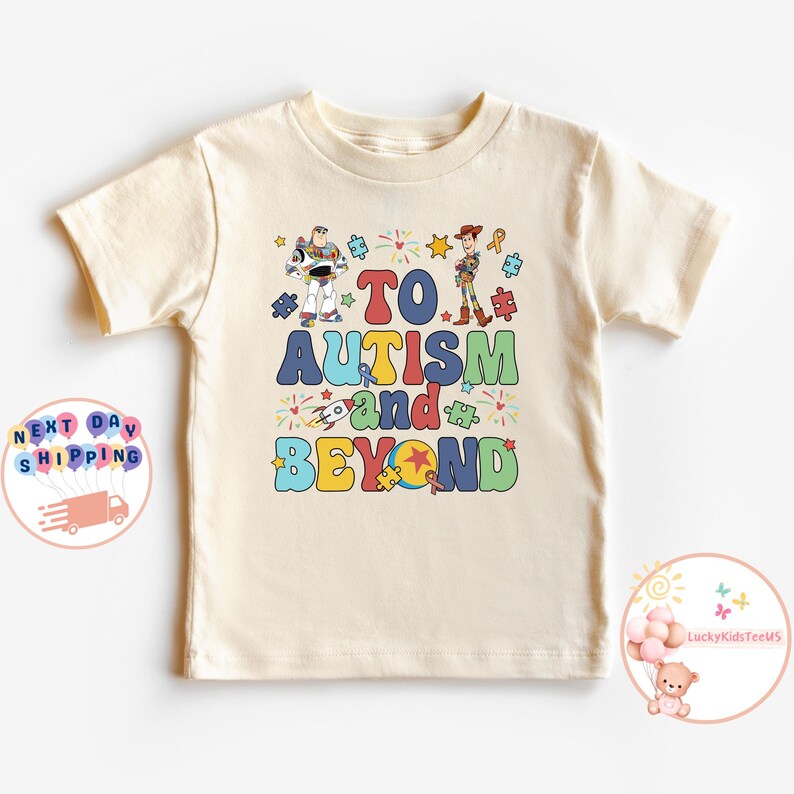 Puede incluir: Una camiseta beige de manga corta con un gr&aacute;fico colorido de los personajes Buzz Lightyear y Woody de la franquicia Toy Story. El gr&aacute;fico incluye el texto "To Autism and Beyond" con piezas de rompecabezas, estrellas y fuegos artificiales.