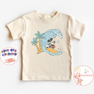 Puede incluir: Una camiseta de color crema con un gr&aacute;fico de Mickey Mouse surfeando en una ola con palmeras en el fondo.
