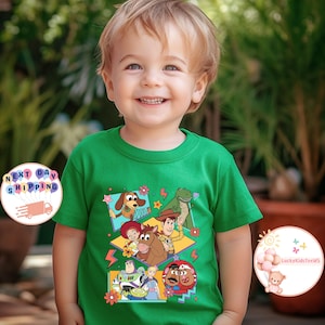 Op de afbeelding: Groen T-shirt met een kleurrijke grafische print van personages uit de Toy Story-filmfranchise. De personages zijn Woody, Buzz Lightyear, Jessie, Rex en Slinky Dog. De grafiek is in retro-stijl met geometrische vormen en bliksemschichten.