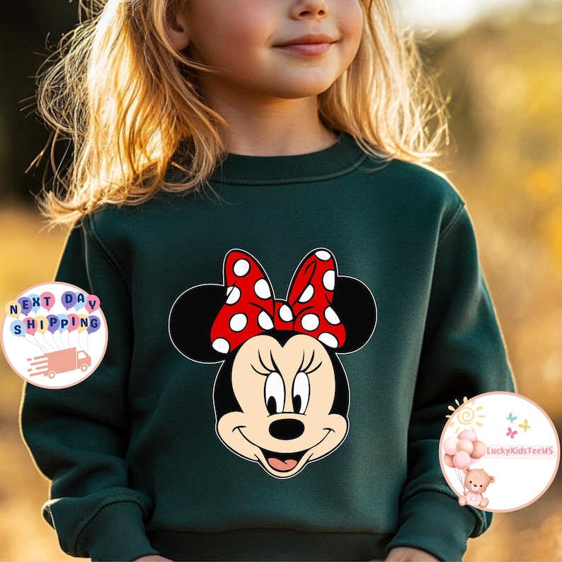 May include: A dark green sweatshirt with a Minnie Mouse design. The graphic depicts Minnie's face, complete with a red bow adorned with white polka dots. The sweatshirt has a crew neck.