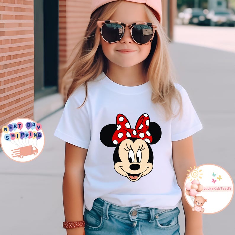 May include: White t-shirt with a Minnie Mouse graphic, featuring a red polka dot bow. The design is centered on the front. The child is wearing blue jeans and sunglasses.