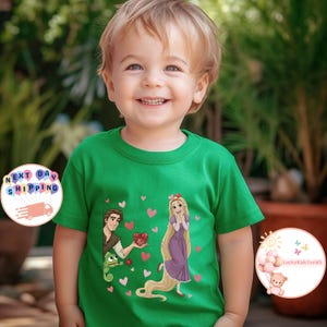 May include: A green t-shirt featuring a cartoon illustration of a princess and a prince, surrounded by pink hearts. The prince is holding a heart-shaped gift. The shirt has a round neck and short sleeves. The text "LuckyKidsTeeUS" is visible.