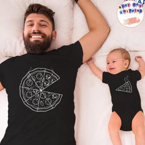 May include: Black t-shirts with a white pizza slice design. One shirt features a full pizza, the other a single slice. The shirts are worn by an adult and a baby, lying on a white surface.