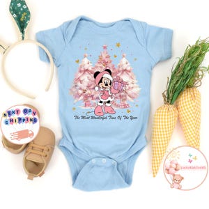 May include: A light blue baby onesie featuring a Minnie Mouse graphic holding a gift in front of pink Christmas trees. The text "The Most Wonderful Time Of The Year" is printed below the graphic. The onesie has short sleeves.