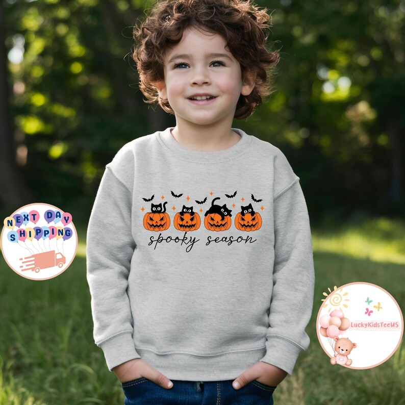 May include: A light gray sweatshirt featuring a Halloween design. The design includes four orange pumpkins with black cat faces, black bats, and the words "spooky season" in black script. The sweatshirt is worn by a child.
