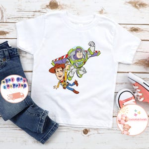 May include: White t-shirt featuring a colorful graphic of Buzz Lightyear and Woody from Toy Story. Buzz is in a flying pose, and Woody is falling. The shirt is paired with blue jeans and pink sneakers. The shirt is likely for children.
