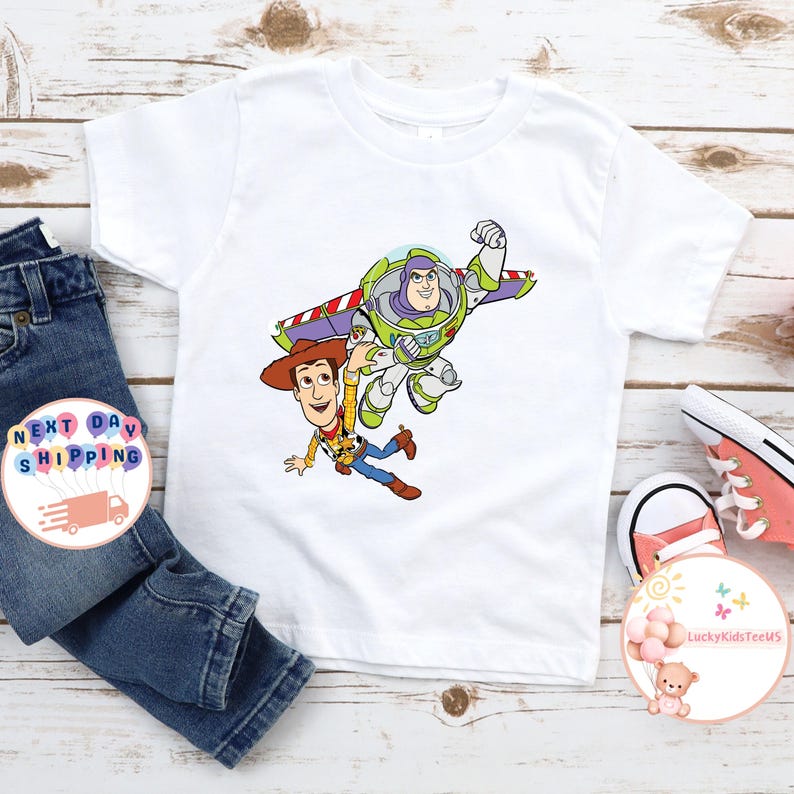 May include: White t-shirt featuring a colorful graphic of Buzz Lightyear and Woody from Toy Story. Buzz is in a flying pose, and Woody is falling. The shirt is paired with blue jeans and pink sneakers. The shirt is likely for children.