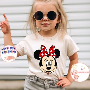 May include: A cream t-shirt with a Minnie Mouse graphic, featuring a red polka-dot bow. The image shows a child wearing the shirt and sunglasses. The shirt is paired with grey jeans. The text "LuckyKidsTeeUS" is visible.
