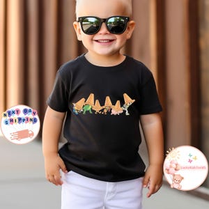 May include: A black t-shirt featuring a colorful graphic of cartoon characters wearing orange hats. The child in the image is wearing black sunglasses and white shorts. The shirt design includes the text "LuckyKidsTeeUS".
