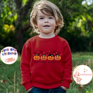 May include: A red sweatshirt featuring a Halloween design. The design includes four jack-o'-lanterns with cat faces, bats, and the words "spooky season" in a script font. The sweatshirt is worn by a young child.