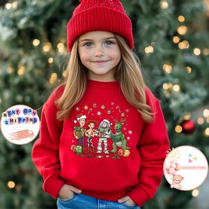 May include: A red sweatshirt with a Christmas-themed design featuring cartoon characters such as Woody, Buzz Lightyear, and Rex, along with festive ornaments. The sweatshirt is worn by a young person wearing a red beanie. The background is a blurred Christmas tree.
