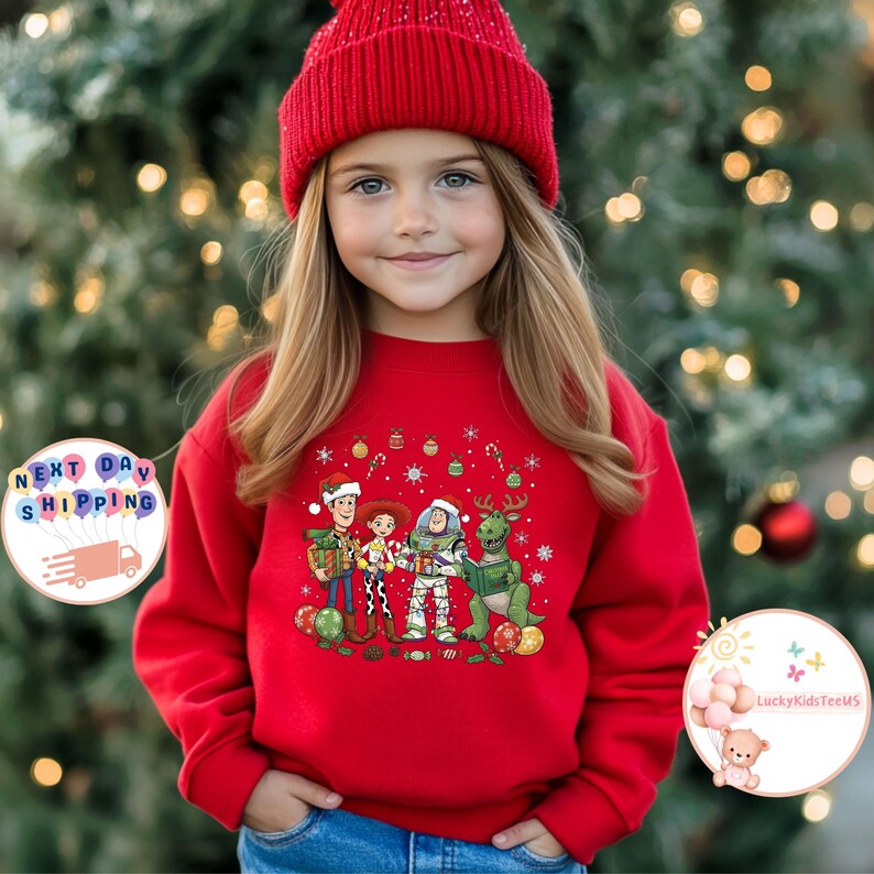 May include: A red sweatshirt with a Christmas-themed design featuring cartoon characters such as Woody, Buzz Lightyear, and Rex, along with festive ornaments. The sweatshirt is worn by a young person wearing a red beanie. The background is a blurred Christmas tree.