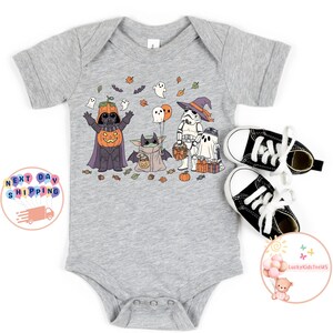 以下が含まれることがあります： ハロウィーンをテーマにした、スターウォーズのキャラクターが仮装したデザインのグレーのベビーボディスーツ。デザインには、カボチャ姿のダース・ベイダー、ストームトルーパー、ベビーヨーダが含まれています。白い靴紐が付いた黒いキャンバススニーカーが隣にあります。