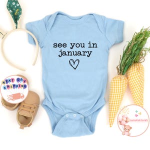 May include: A light blue baby onesie with the text "see you in january" and a small heart.