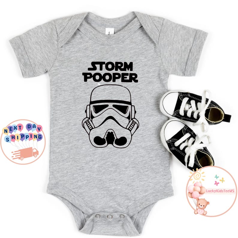 May include: A gray baby onesie with a black graphic of a Star Wars Stormtrooper helmet and the text "Storm Pooper".