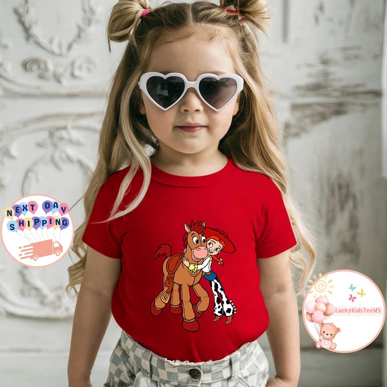 May include: A red t-shirt featuring a cartoon image of a horse and a cowgirl hugging. The shirt is worn by a young child wearing heart-shaped sunglasses. The child has blonde hair styled in pigtails.