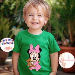 May include: A green t-shirt featuring a Minnie Mouse graphic. Minnie is depicted in pink with a bow, holding a teddy bear. The shirt is worn by a young child.