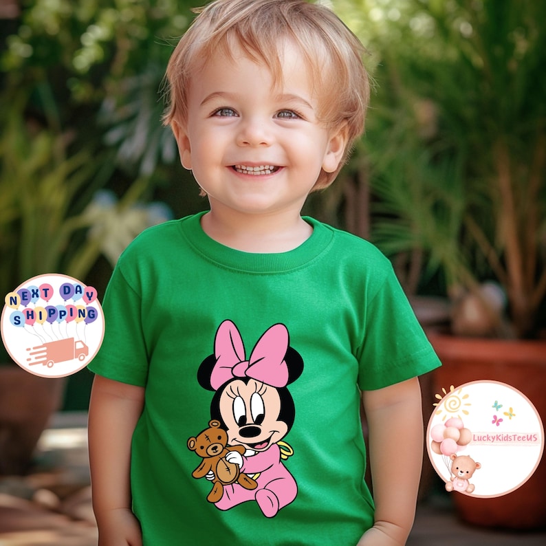 May include: A green t-shirt featuring a Minnie Mouse graphic. Minnie is depicted in pink with a bow, holding a teddy bear. The shirt is worn by a young child.