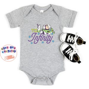 May include: A gray baby onesie featuring a graphic of Buzz Lightyear with the phrase "To Infinity" in purple script. The onesie is paired with black and white baby sneakers. The image has a white background.