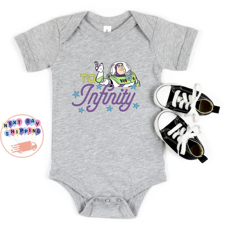 May include: A gray baby onesie featuring a graphic of Buzz Lightyear with the phrase "To Infinity" in purple script. The onesie is paired with black and white baby sneakers. The image has a white background.