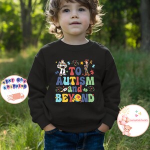 Puede incluir: Una camiseta negra de manga larga con un gr&aacute;fico de los personajes de Toy Story, Buzz Lightyear y Woody. El gr&aacute;fico incluye el texto "To Autism and Beyond" con piezas de rompecabezas y fuegos artificiales.