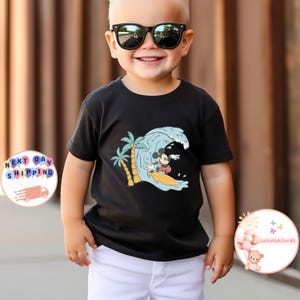 Puede incluir: Camiseta negra con un gr&aacute;fico de Mickey Mouse surfeando en una ola con palmeras en el fondo.