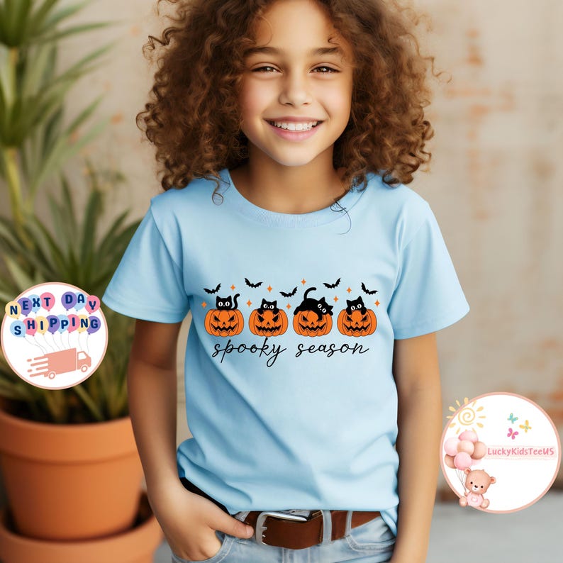 May include: A light blue t-shirt featuring a Halloween design. The design includes four orange jack-o'-lanterns with black cat faces, black bats, and the words "spooky season" in a script font. The shirt is worn by a young person.
